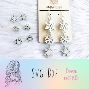 May include: White snowflake earrings with three snowflakes on each earring. The earrings are displayed on a wooden card with the text "Shelby Designing". The image also includes a close-up of the snowflake earrings and the text "SVG DXF laser cut file".