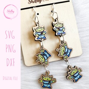 May include: Wooden dangle earrings featuring green aliens in blue uniforms. The earrings are connected by silver-colored chains and hang from silver hooks. The earrings are displayed on a wooden surface. The text "SVG, PNG, DXF, DIGITAL FILE" is on the left.