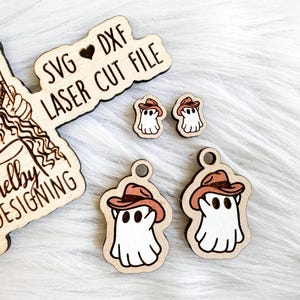 K&ouml;nnte beinhalten: Ein Set aus vier lasergeschnittenen Holz-Geister-Anh&auml;ngern. Die Geister tragen Cowboyh&uuml;te und haben ein verspieltes Design. Die Anh&auml;nger sind perfekt f&uuml;r Halloween-Dekorationen oder Bastelprojekte. Das Bild zeigt auch ein Schild mit der Aufschrift "SVG &hearts; DXF LASER CUT FILE Shelby DESIGNING".