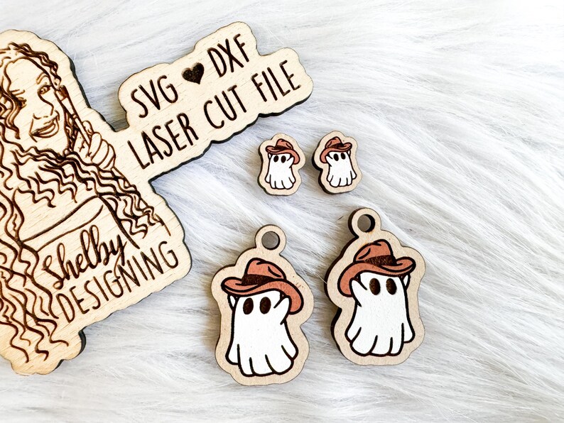 K&ouml;nnte beinhalten: Ein Set aus vier lasergeschnittenen Holz-Geister-Anh&auml;ngern. Die Geister tragen Cowboyh&uuml;te und haben ein verspieltes Design. Die Anh&auml;nger sind perfekt f&uuml;r Halloween-Dekorationen oder Bastelprojekte. Das Bild zeigt auch ein Schild mit der Aufschrift "SVG &hearts; DXF LASER CUT FILE Shelby DESIGNING".