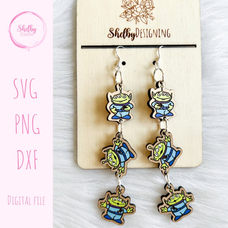May include: Wooden dangle earrings featuring three green alien figures with blue and white accents. The earrings are connected by silver-colored chains and hang from silver hooks. The earrings are displayed on a wooden backing with the text "Shelby Designing".