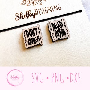 May include: Two wooden earrings with the phrases "DON'T OPEN" and "DEAD INSIDE" in a distressed font. The earrings are on a light wood surface with the name "Shelby Designing" and floral accents. A black line and the text "SVG • PNG • DXF" are also visible.