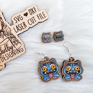 May include: Wooden earrings and studs featuring a cartoon cat design with blue, yellow, and pink accents. The earrings have silver hooks. Also included is a wooden sign that says "SVG DXF LASER CUT FILE" and "Shelby DESIGNING".