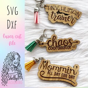 May include: Three wooden keychains with different phrases and tassels. The first keychain says "Tiny Human Trainer" with a pink tassel. The second keychain says "Chaos Coordinator" with a green tassel. The third keychain says "Mommin' All Day Err Day" with a yellow tassel.