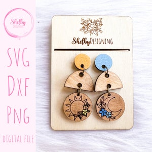 May include: A pair of wooden earrings with a sun and moon design. The earrings are painted in shades of yellow, blue, and brown. The sun has a yellow center and a brown outline with rays. The moon has a blue center and a brown outline with a crescent shape. The earrings are hanging from a wooden display card with the text "ShelbyDESIGNING" printed on it.