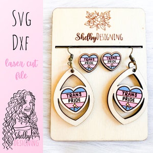 May include: A set of laser cut wooden earrings featuring a transgender pride heart design. The earrings are in the shape of a teardrop with a heart cutout in the center. The heart is painted with the transgender pride colors: light blue, pink, and white. The earrings are displayed on a wooden stand with the text "Shelby DESIGNING" printed on it.