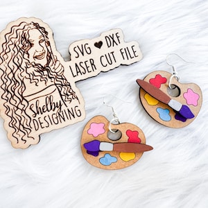 May include: A pair of wooden earrings shaped like paint palettes with paintbrushes. The earrings are brown with colourful paint splatters and a purple paintbrush. The earrings are labelled "SVG ♥ DXF LASER CUT FILE".