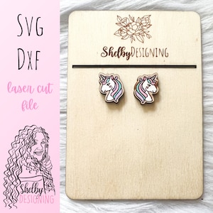 May include: A pair of wooden unicorn stud earrings with pink, blue, and white details. The earrings are displayed on a wooden card with the text "Shelby DESIGNING".