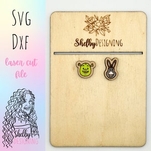 May include: Wooden display board with two cartoon character earrings: Shrek and a donkey. The board has the text "SVG DXF laser cut file" and the name "Shelby DESIGNING". The earrings are on a light wood background.