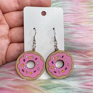 May include: A pair of donut-shaped earrings with pink frosting and colorful sprinkles. The earrings are made of wood and have silver hooks.