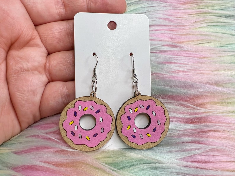 May include: A pair of donut-shaped earrings with pink frosting and colorful sprinkles. The earrings are made of wood and have silver hooks.