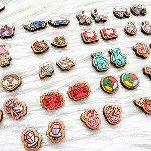 May include: A collection of 20 pairs of wooden earrings featuring nostalgic 90s toys and games. The earrings are painted in bright colors and feature designs such as a Rubik's Cube, a Tamagotchi, a Troll doll, a View-Master, and a game of Operation.