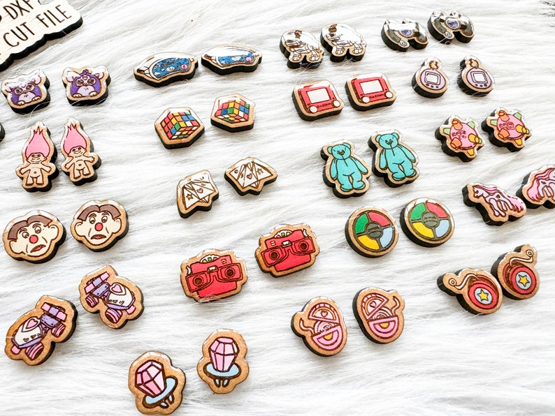 May include: A collection of 20 pairs of wooden earrings featuring nostalgic 90s toys and games. The earrings are painted in bright colors and feature designs such as a Rubik's Cube, a Tamagotchi, a Troll doll, a View-Master, and a game of Operation.