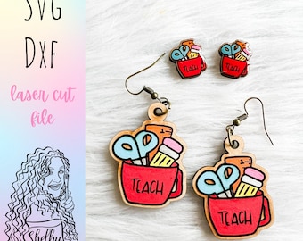 May include: A pair of laser cut wooden earrings featuring a red coffee mug with the word "TEACH" on it. The mug is filled with school supplies, including a pencil, ruler, and scissors. The earrings are a fun and unique way to show your love for teaching.