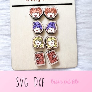 May include: Six pairs of laser cut wooden earrings featuring different characters from the movie Hocus Pocus. The earrings are in the shape of a witch's face with different hair colors and facial expressions. There are also two pairs of earrings in the shape of a closed book with a red cover and a gold eye design.