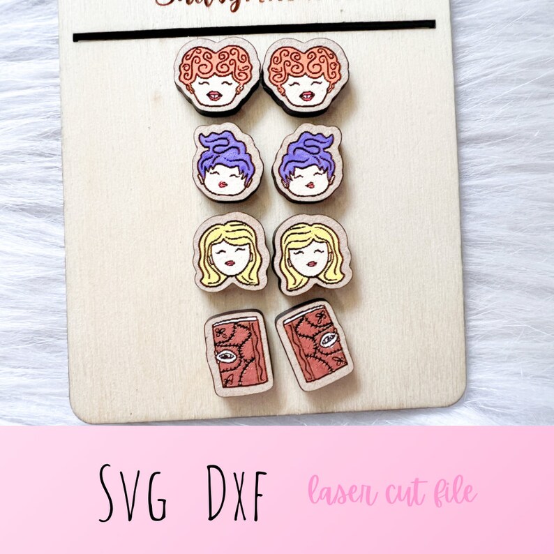 May include: Six pairs of laser cut wooden earrings featuring different characters from the movie Hocus Pocus. The earrings are in the shape of a witch's face with different hair colors and facial expressions. There are also two pairs of earrings in the shape of a closed book with a red cover and a gold eye design.