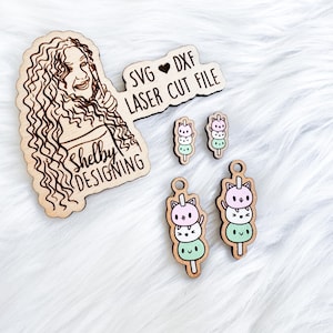 May include: A set of laser cut wood earrings featuring a stack of three mochi with cute cat faces. The earrings are labeled "SVG ♥ DXF LASER CUT FILE" and "Shelby Designing".