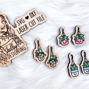 May include: A set of five laser cut wooden earrings featuring hanging potted plants with cute faces. The earrings are available as SVG and DXF files for digital download. The text "SVG ♥ DXF LASER CUT FILE" and "Shelby Designing" are also included in the image.