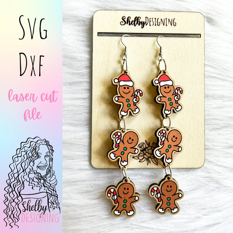May include: A pair of gingerbread man dangle earrings with red, green, and white details. The earrings are made of wood and have a silver hook.