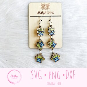 May include: Wooden dangle earrings featuring the Little Green Men from Toy Story. Each earring has three alien figures in blue and green, connected by silver-tone metal. The earrings hang from silver hooks. The background is a light wood and white fur.