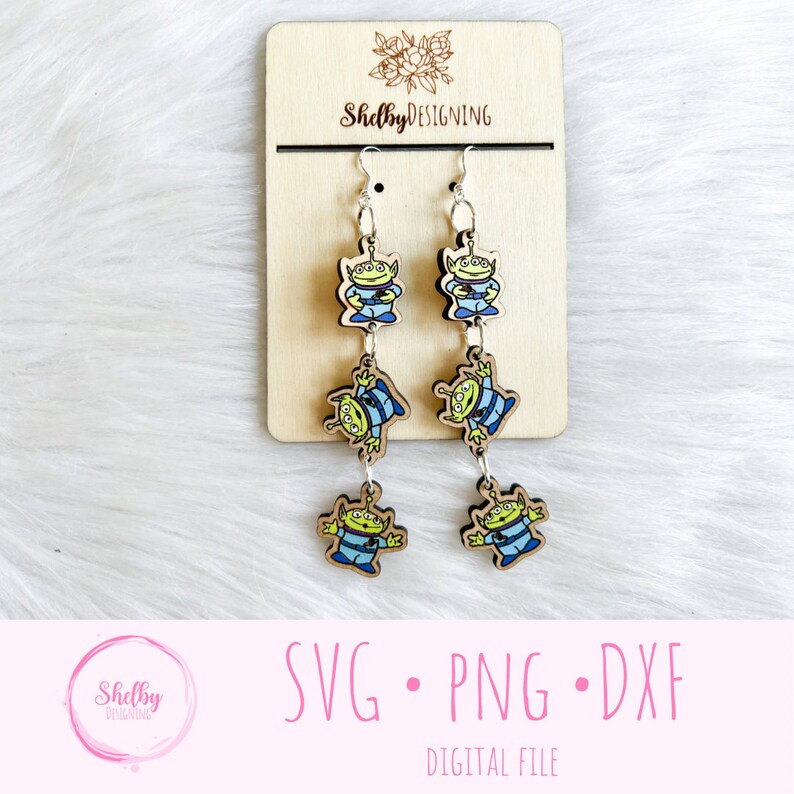 May include: Wooden dangle earrings featuring the Little Green Men from Toy Story. Each earring has three alien figures in blue and green, connected by silver-tone metal. The earrings hang from silver hooks. The background is a light wood and white fur.