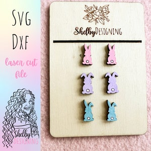 Pode incluir: Um conjunto de seis brincos de coelho de madeira cortados a laser nas cores rosa, roxo e azul. Os brincos são exibidos em uma placa de madeira com o texto "Shelby DESIGNING" no topo.