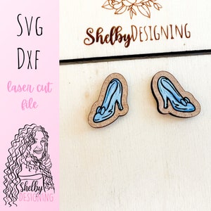 May include: Two wooden Cinderella-style high heel shoe cutouts with blue painted details. The shoes are on a light wood surface. The image also includes the text "SVG DXF laser cut file" and the name "Shelby Designing".
