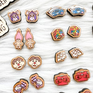 May include: A collection of 16 pairs of wooden earrings featuring nostalgic 80s and 90s pop culture icons. The earrings are painted in bright colors and feature designs like a Rubik's Cube, a troll doll, a Care Bear, a cassette tape, and a vintage camera.