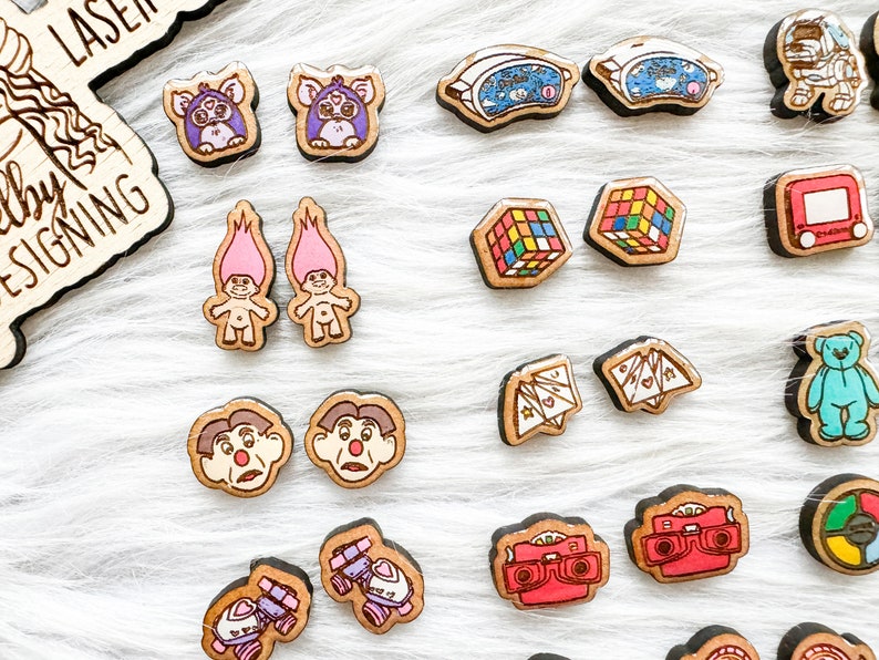 May include: A collection of 16 pairs of wooden earrings featuring nostalgic 80s and 90s pop culture icons. The earrings are painted in bright colors and feature designs like a Rubik's Cube, a troll doll, a Care Bear, a cassette tape, and a vintage camera.