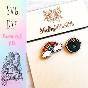 May include: Laser cut wooden earrings featuring a rainbow and a pot of gold. The earrings are on a wooden display card with the text "Shelby DESIGNING".