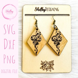 May include: A pair of wooden earrings with a snake design and a diamond shape. The earrings are on a wooden display stand with the text "Shelby Designing" on it.
