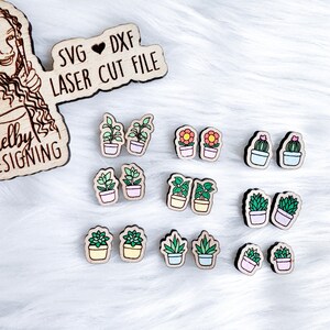May include: A set of 10 pairs of laser cut wooden earrings featuring different potted plants, including succulents, cacti, and flowers. The earrings are painted in various colors, including green, pink, yellow, and orange. The earrings are arranged in two rows on a white fuzzy surface. The text "SVG ♥ DXF LASER CUT FILE" is visible in the top left corner of the image.