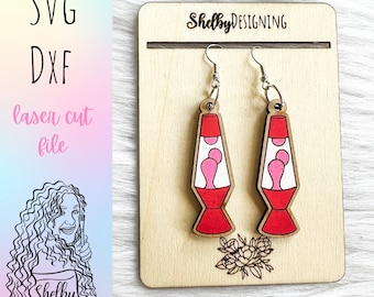 May include: A pair of laser cut wooden earrings shaped like lava lamps. The lamps are red with pink lava inside. The earrings are hanging on a wooden display card with the text "ShelbyDESIGNING" printed on it.