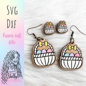 May include: A pair of laser cut wooden earrings shaped like Easter baskets filled with colorful eggs. The earrings have a yellow bow on top and are perfect for spring.