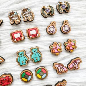 May include: A collection of 18 colorful wooden earrings featuring various nostalgic 90s and early 2000s pop culture icons. The earrings include a Tamagotchi, a Game Boy, a Rubik's Cube, a teddy bear, a Game & Watch, and a Nintendo character.