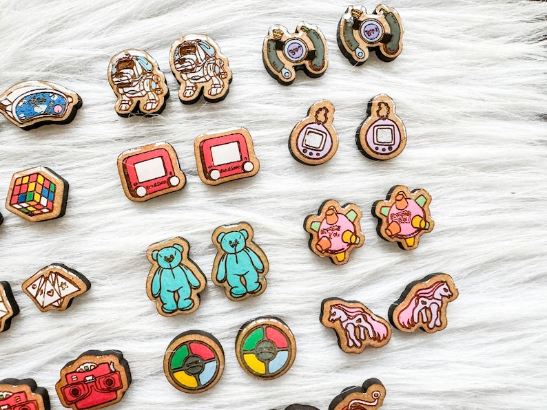 May include: A collection of 18 colorful wooden earrings featuring various nostalgic 90s and early 2000s pop culture icons. The earrings include a Tamagotchi, a Game Boy, a Rubik's Cube, a teddy bear, a Game & Watch, and a Nintendo character.