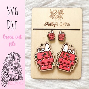 May include: Wooden earrings featuring Snoopy in his doghouse, adorned with Christmas lights. The earrings are displayed on a wooden card with the text "Shelby Designing" and "SVG DXF laser cut file".