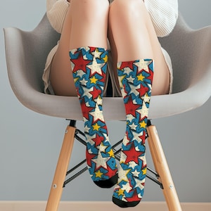 May include: A pair of knee-high socks featuring a repeating pattern of red, white, and yellow stars on a blue background. The socks have black toes and heels. The socks are worn by a person sitting in a gray chair.