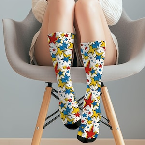May include: A pair of knee-high socks with a white background and a repeating pattern of red, yellow, and blue stars. The socks have black toes and heels. The person is sitting in a gray chair.