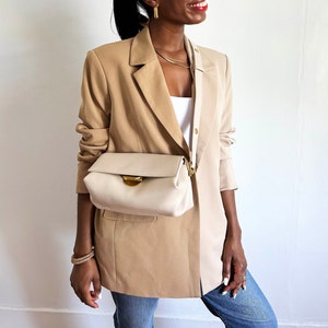 May include: A tan blazer with a cream-colored crossbody bag featuring a gold clasp. The bag is worn over a white top. The outfit is completed with blue jeans and gold jewelry, creating a stylish, everyday look.