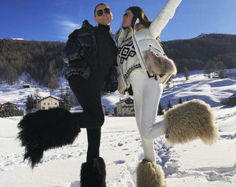 May include: Two people pose in the snow, wearing winter attire. One wears a black puffer jacket, leggings, and black furry boots. The other wears a white jacket, ski pants, and beige furry boots. One holds a champagne bottle.