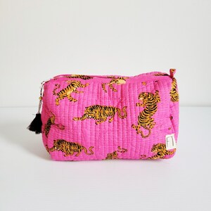 Op de afbeelding: Een gewatteerde, rechthoekige make-up tas in een levendige roze tint. De tas is versierd met een herhalend patroon van oranje en zwarte tijgerillustraties. Een zwarte kwast hangt aan de ritssluiting. De tas staat op een wit oppervlak.