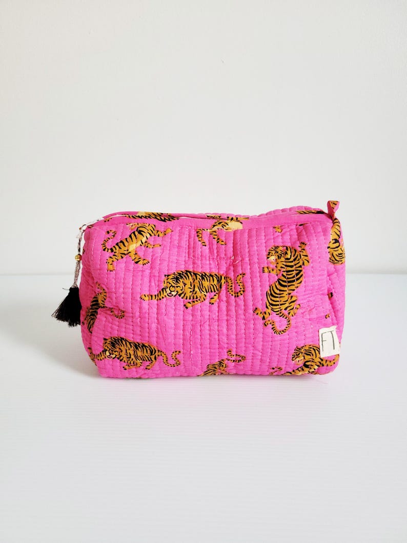 Op de afbeelding: Een gewatteerde, rechthoekige make-up tas in een levendige roze tint. De tas is versierd met een herhalend patroon van oranje en zwarte tijgerillustraties. Een zwarte kwast hangt aan de ritssluiting. De tas staat op een wit oppervlak.