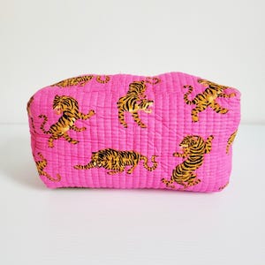 Op de afbeelding: Een gewatteerde, rechthoekige make-uptas in een levendige roze tint. De tas is versierd met een herhalend patroon van gestileerde tijgers in zwart en geel. De tas is ongeveer 25 cm lang.