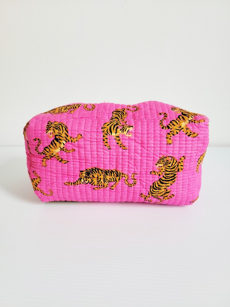 Op de afbeelding: Een gewatteerde, rechthoekige make-uptas in een levendige roze tint. De tas is versierd met een herhalend patroon van gestileerde tijgers in zwart en geel. De tas is ongeveer 25 cm lang.