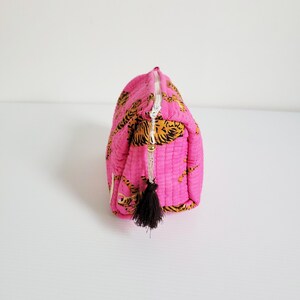 Op de afbeelding: Een gewatteerde, roze make-uptas met een goud-zwarte tijgerprint. De tas heeft een witte rits en een zwarte kwast. De tas is rechthoekig en heeft een afgeronde bovenkant.