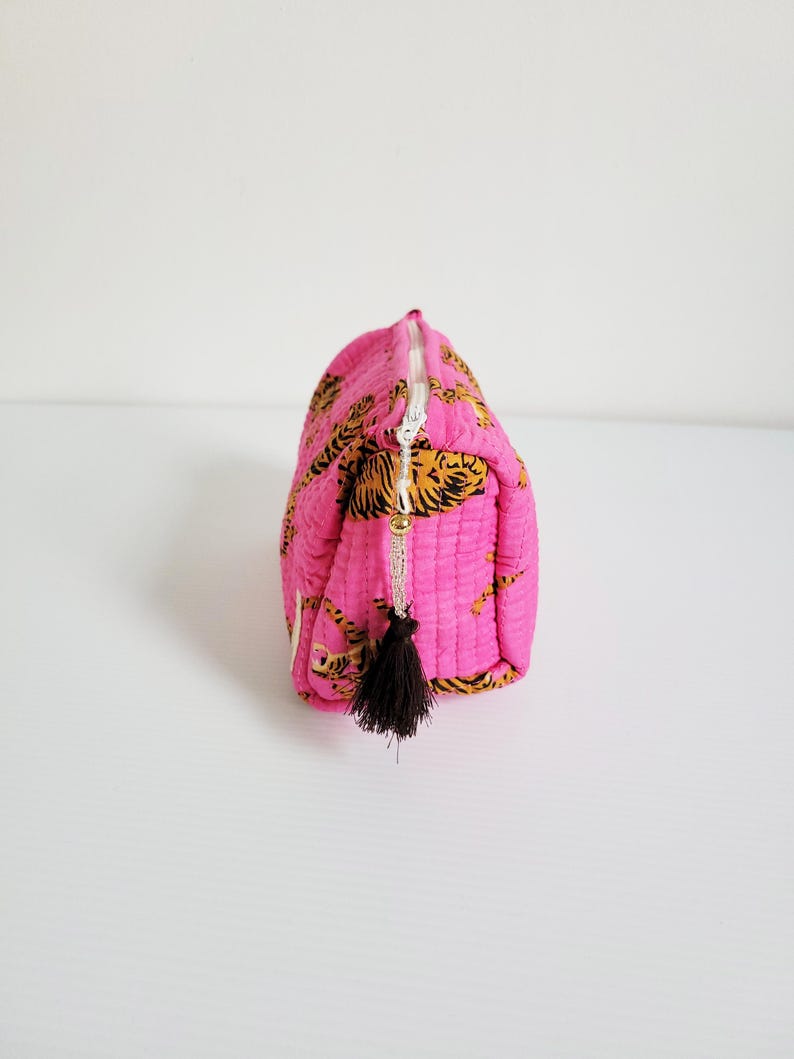 Op de afbeelding: Een gewatteerde, roze make-uptas met een goud-zwarte tijgerprint. De tas heeft een witte rits en een zwarte kwast. De tas is rechthoekig en heeft een afgeronde bovenkant.