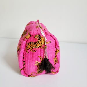 Op de afbeelding: Een gewatteerde, roze make-uptas met een herhalend patroon van oranje en zwarte tijgers. De tas heeft een zwarte kwast en een witte rits. De tas staat op een wit oppervlak.