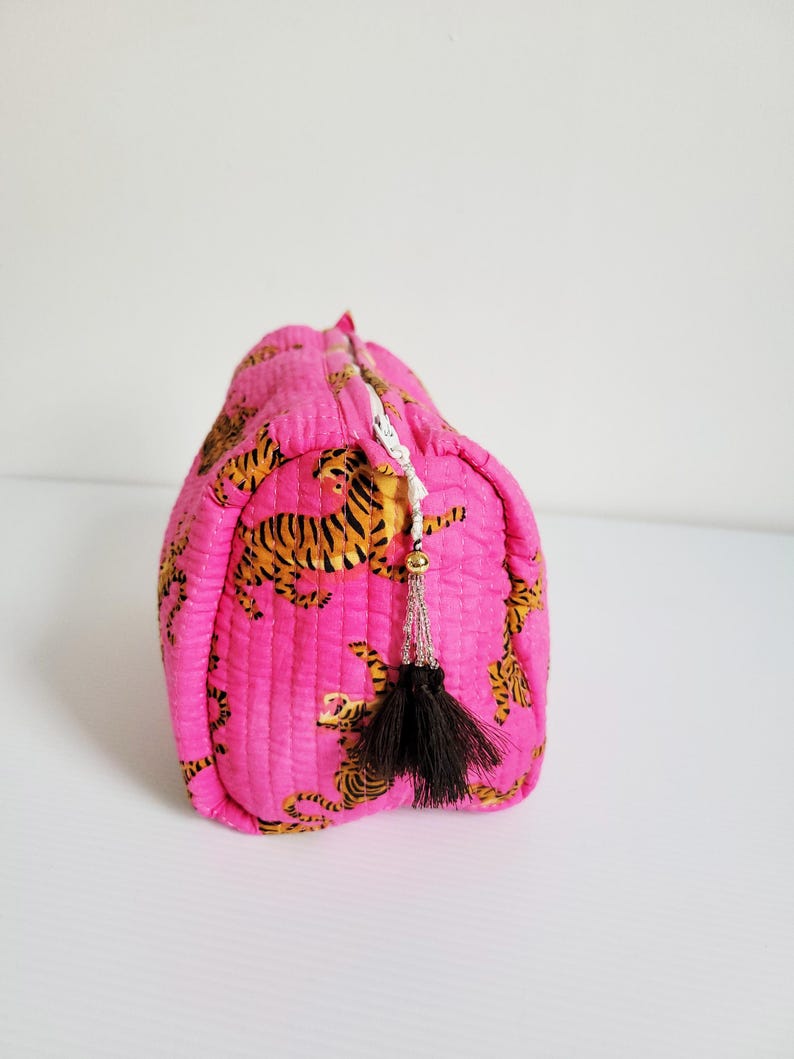 Op de afbeelding: Een gewatteerde, roze make-uptas met een herhalend patroon van oranje en zwarte tijgers. De tas heeft een zwarte kwast en een witte rits. De tas staat op een wit oppervlak.