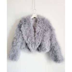 Könnte beinhalten: Eine graue Federjacke mit flauschiger Textur. Die Jacke hängt an einem weißen Kleiderbügel vor einem weißen Hintergrund.